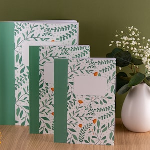 Puede incluir: Conjunto de tres cuadernos verdes y blancos con un patrón floral. Los cuadernos tienen una encuadernación blanca y un borde verde. El patrón presenta pequeñas hojas verdes y flores naranjas.