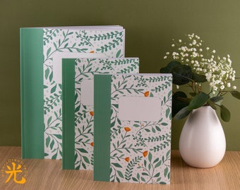 May include: Set of three green and white notebooks with a floral pattern. The notebooks have a white spine and a green border. The pattern features small green leaves and orange flowers.