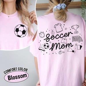 K&ouml;nnte beinhalten: Hellrosa T-Shirt mit einem schwarz-wei&szlig;en Fu&szlig;ball-Grafik auf der Vorderseite. Auf der R&uuml;ckseite des Shirts steht "Soccer Mom" mit Fu&szlig;ball-Themen-Kritzeleien. Das Shirt ist mit "Comfort Color Blossom" gekennzeichnet.