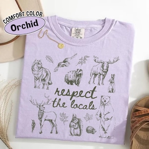 May include: A lilac Comfort Colors t-shirt with a graphic of woodland creatures and the text "respect the locals." The design includes a wolf, moose, bear, and other wildlife.