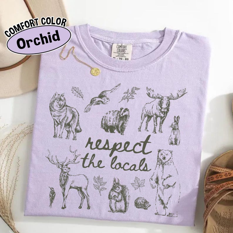 May include: A lilac Comfort Colors t-shirt with a graphic of woodland creatures and the text "respect the locals." The design includes a wolf, moose, bear, and other wildlife.