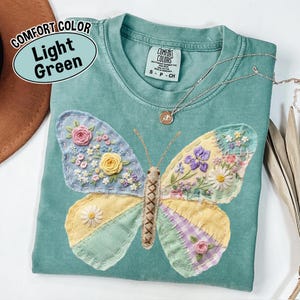 May include: Light green Comfort Colors t-shirt with an embroidered butterfly design. The butterfly has yellow, blue, and purple wings with floral details. A silver necklace with a small pendant rests on the shirt.