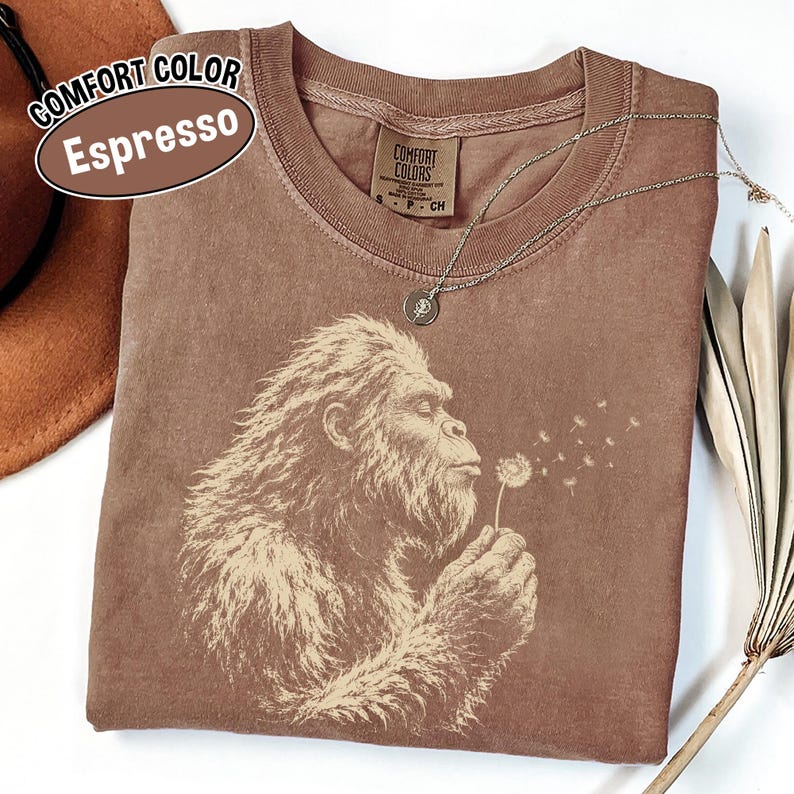 Puede incluir: Una camiseta de cuello redondo color espresso con un estampado gr&aacute;fico de una criatura peluda soplando un diente de le&oacute;n. La camiseta tiene una etiqueta de la marca Comfort Colors en el cuello interior. El dise&ntilde;o muestra a la criatura de perfil.