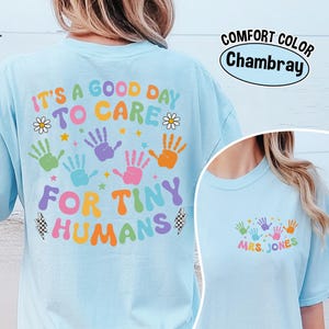 Puede incluir: Camiseta azul claro con el texto "IT'S A GOOD DAY TO CARE FOR TINY HUMANS" en letras coloridas y huellas de manos. La camiseta tambi&eacute;n tiene el texto "MRS. JONES" en la parte delantera. La camiseta es Comfort Color Chambray.