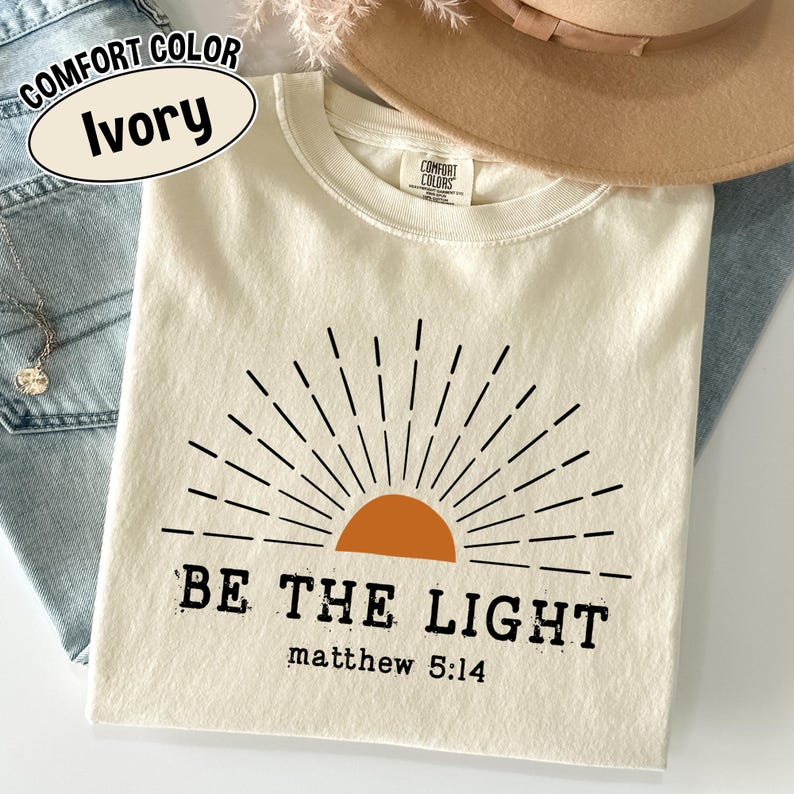 May include: Ivory-colored t-shirt with a graphic of a rising sun and the text "BE THE LIGHT" and "matthew 5:14". The shirt is folded and displayed with a hat and jeans. The shirt is labeled "Comfort Color".