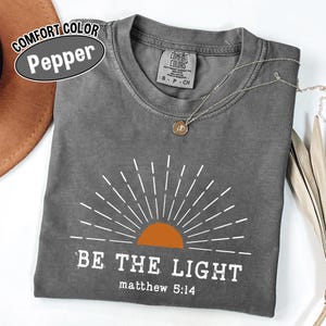 May include: A gray Comfort Colors t-shirt with the text "BE THE LIGHT" and "matthew 5:14" printed in white. The design includes a sun graphic with an orange center and white rays. A silver necklace with a pendant is visible.