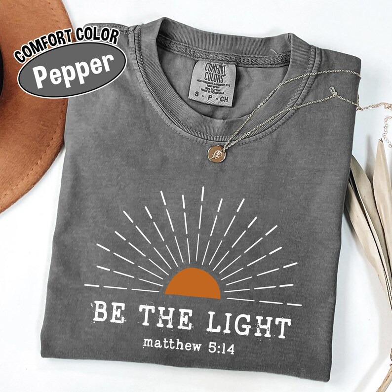 May include: A gray Comfort Colors t-shirt with the text "BE THE LIGHT" and "matthew 5:14" printed in white. The design includes a sun graphic with an orange center and white rays. A silver necklace with a pendant is visible.