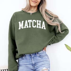 May include: A green sweatshirt with the word "MATCHA" printed in white on the front. The sweatshirt is made of a soft, comfortable material and has a relaxed fit.