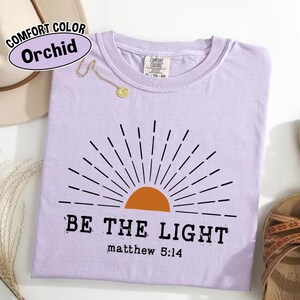 May include: A light purple t-shirt with the text "BE THE LIGHT" and "matthew 5:14" printed in black. The shirt features a graphic of a rising sun in orange and black. The shirt is labeled "Comfort Color Orchid".