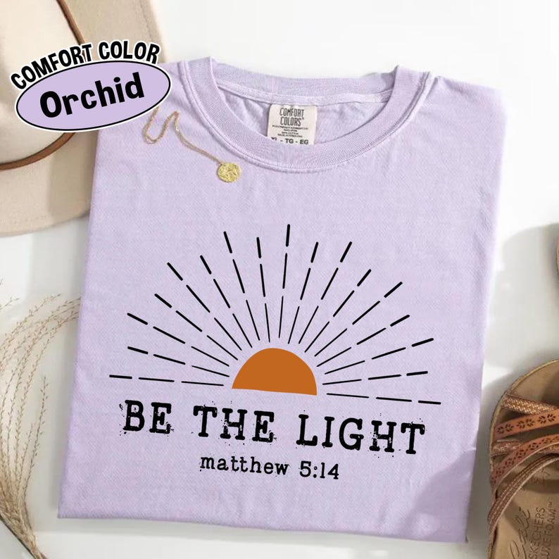 May include: A light purple t-shirt with the text "BE THE LIGHT" and "matthew 5:14" printed in black. The shirt features a graphic of a rising sun in orange and black. The shirt is labeled "Comfort Color Orchid".