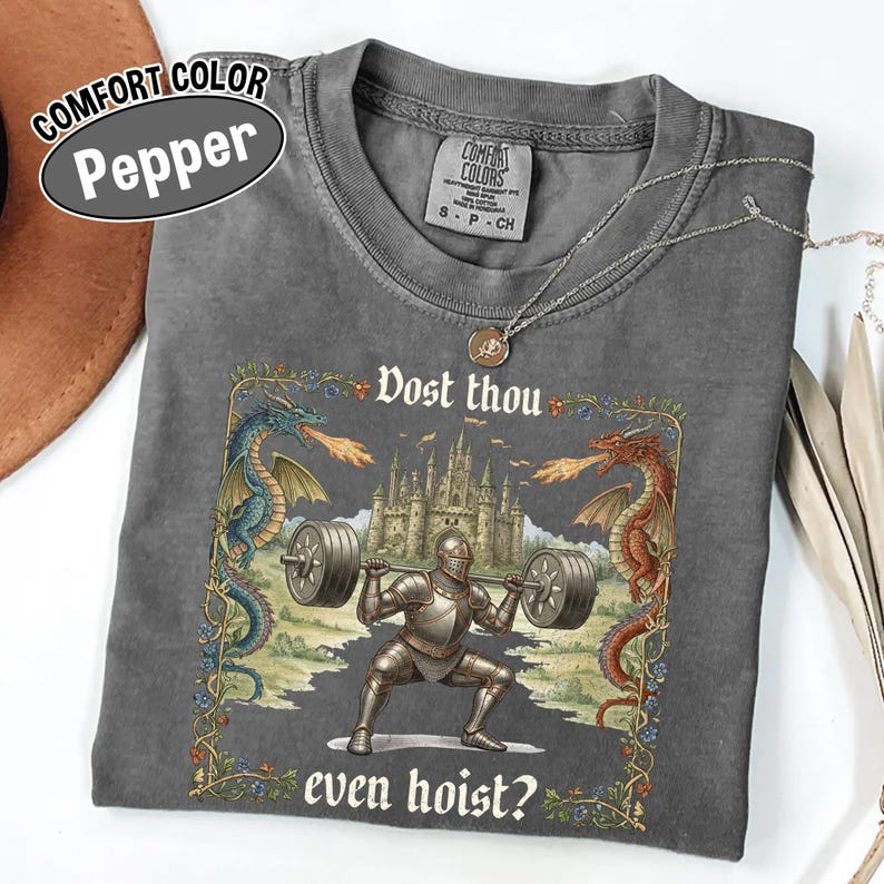 May include: A gray Comfort Colors t-shirt with a graphic of a knight lifting weights, flanked by dragons. The text reads "Dost thou even hoist?" and "Comfort Color Pepper". A silver necklace is draped on the shirt.