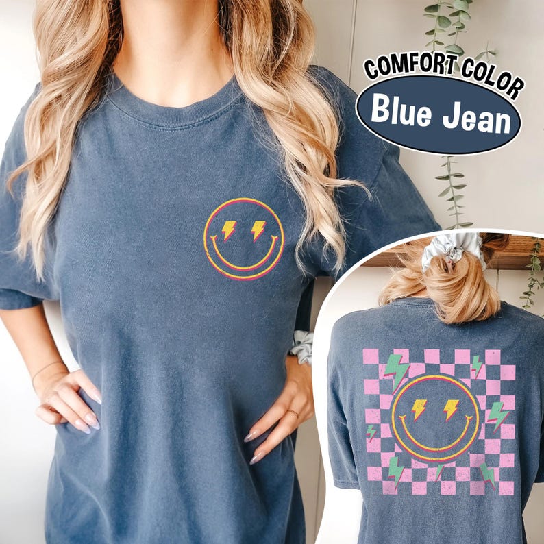 May include: Blue jean colored t-shirt with a smiley face and lightning bolt graphic. The front of the shirt features a small smiley face with lightning bolts. The back of the shirt has a larger smiley face design with a checkered pattern and lightning bolts. The text "COMFORT COLOR Blue Jean" is also visible.