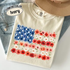 Peut inclure: Un t-shirt à manches courtes ivoire avec un motif représentant le drapeau américain composé de fleurs rouges, blanches et bleues. La section bleue inclut des fleurs blanches en forme d'étoile. L'étiquette Comfort Colors et le texte COMFORT COLOR Ivory.