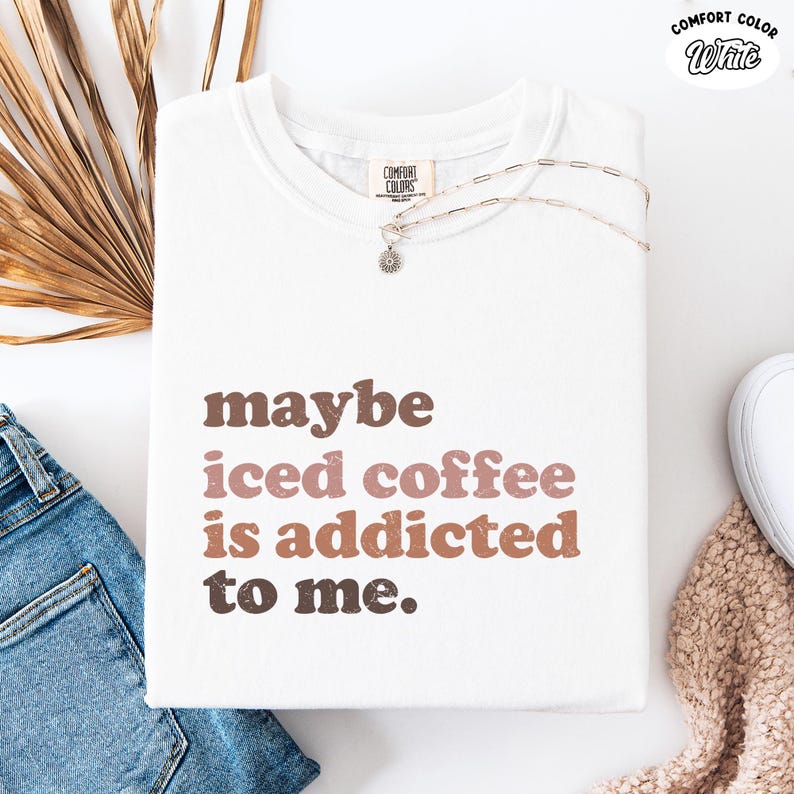 Peut inclure: T-shirt blanc Comfort Colors avec le texte "maybe iced coffee is addicted to me" en lettres marron et roses. Un collier argent&eacute; avec un pendentif en forme de fleur est sur le t-shirt. Le t-shirt est pli&eacute; sur une surface blanche.