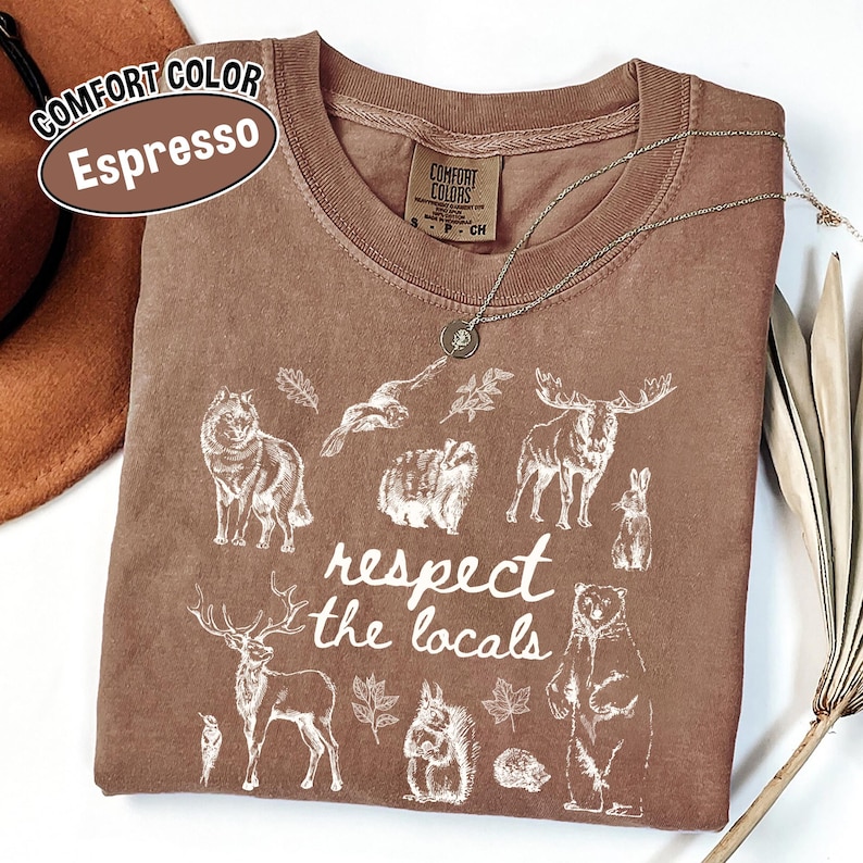 May include: Espresso-coloured Comfort Colors t-shirt with a crew neck and the phrase "respect the locals" in white. The shirt features white illustrations of various animals, including a wolf, moose, and bear. A silver necklace is draped across the shirt.