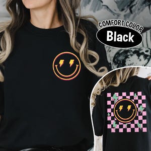 May include: Black crew neck t-shirt with a retro smiley face design featuring lightning bolts. The shirt has a distressed look and the text "COMFORT COLOR Black" is visible. The back of the shirt has a checkered pattern with a smiley face and lightning bolts.