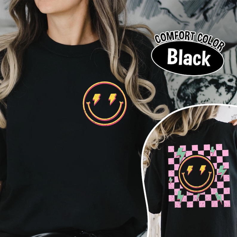 May include: Black crew neck t-shirt with a retro smiley face design featuring lightning bolts. The shirt has a distressed look and the text "COMFORT COLOR Black" is visible. The back of the shirt has a checkered pattern with a smiley face and lightning bolts.