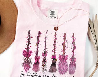 May include: A light pink Comfort Colors t-shirt featuring a graphic of six decorative witch brooms in shades of pink and brown. The text "In October We Wear Pink" is printed below the brooms. A gold necklace is draped across the shirt.