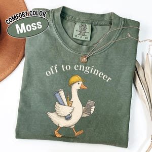 May include: A moss green t-shirt featuring a graphic of a white goose wearing a yellow hard hat, carrying blue rolled blueprints and a pencil under one wing, and holding a calculator in the other. The text reads off to engineer.