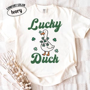 Puede incluir: Camiseta color marfil con un dise&ntilde;o verde de "Lucky Duck". El gr&aacute;fico muestra un pato blanco adornado con tr&eacute;boles. Tr&eacute;boles adicionales est&aacute;n dispersos alrededor del pato. La camiseta tiene una etiqueta Comfort Color.