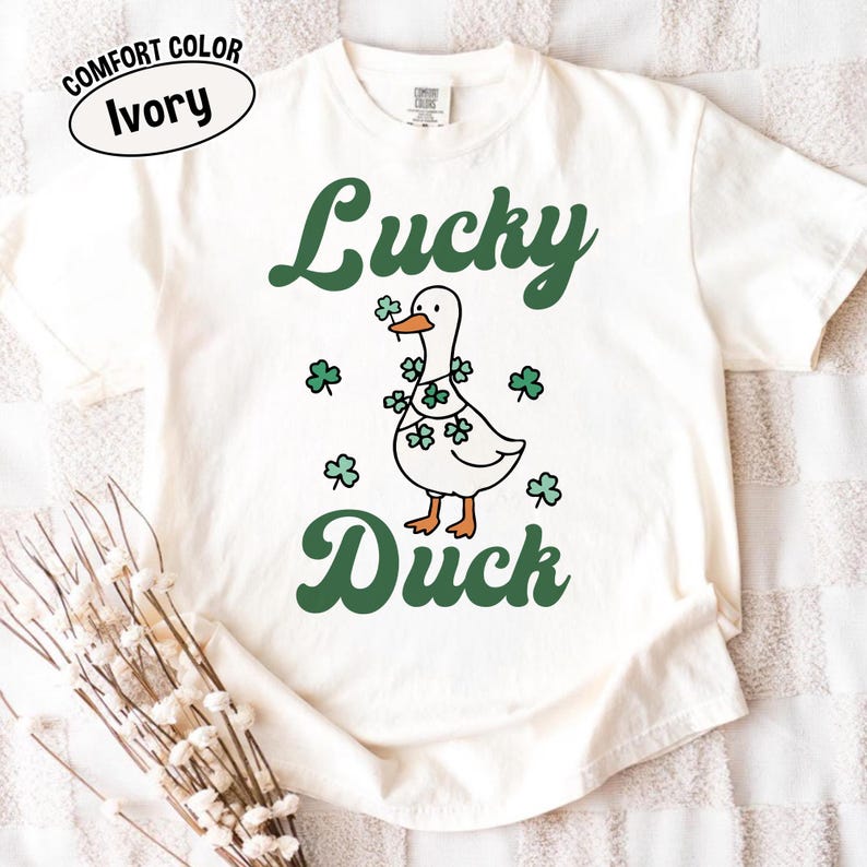 Puede incluir: Camiseta color marfil con un dise&ntilde;o verde de "Lucky Duck". El gr&aacute;fico muestra un pato blanco adornado con tr&eacute;boles. Tr&eacute;boles adicionales est&aacute;n dispersos alrededor del pato. La camiseta tiene una etiqueta Comfort Color.