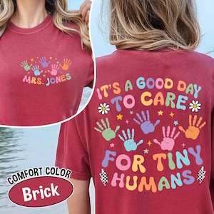 Puede incluir: Camiseta color ladrillo con el texto "MRS. JONES" y huellas de manos coloridas en la parte delantera. La parte trasera de la camiseta dice "IT'S A GOOD DAY TO CARE FOR TINY HUMANS" con huellas de manos y elementos decorativos.