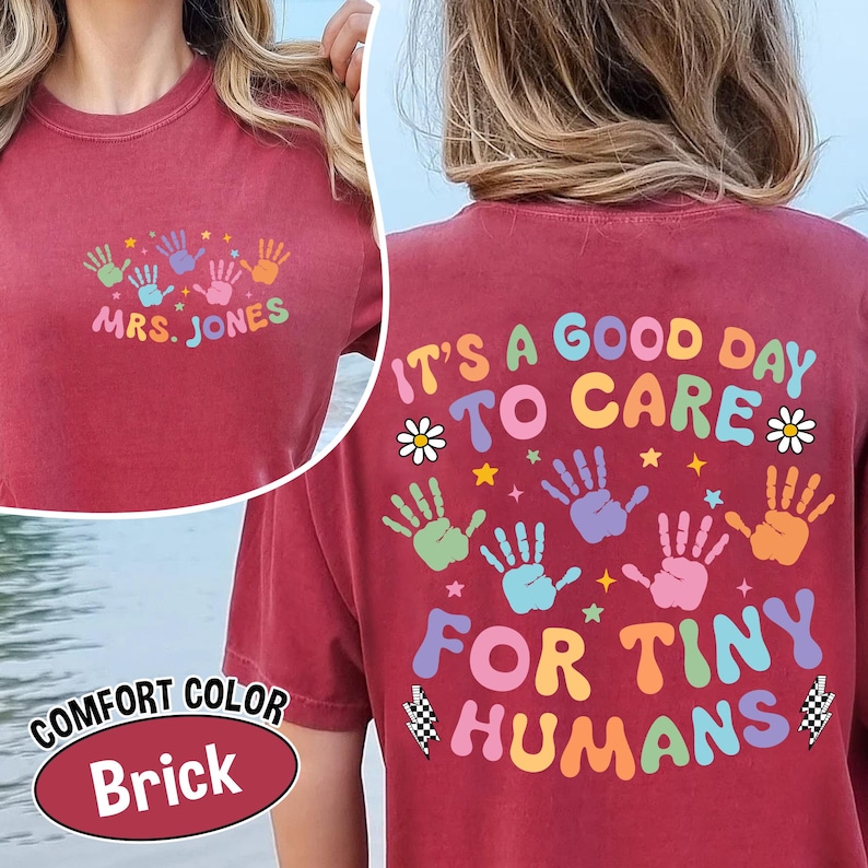 Puede incluir: Camiseta color ladrillo con el texto "MRS. JONES" y huellas de manos coloridas en la parte delantera. La parte trasera de la camiseta dice "IT'S A GOOD DAY TO CARE FOR TINY HUMANS" con huellas de manos y elementos decorativos.