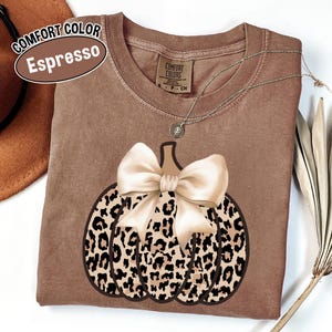 Peut inclure: T-shirt Comfort Colors couleur Espresso avec un motif de citrouille à imprimé léopard et un nœud crème. Un collier argenté avec un pendentif est drapé sur le t-shirt. Le t-shirt est à col rond.