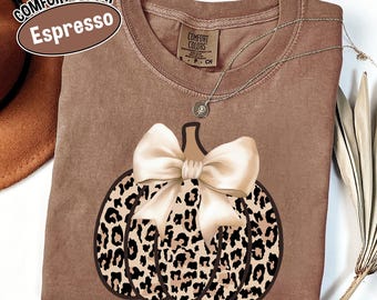 May include: Espresso-colored Comfort Colors t-shirt featuring a pumpkin design with a leopard print pattern and a cream-colored bow. A silver necklace with a pendant is draped over the shirt. The shirt is a crew neck.