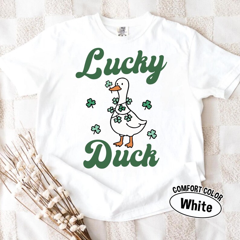 Puede incluir: Camiseta blanca Comfort Color con un gr&aacute;fico verde "Lucky Duck". El gr&aacute;fico presenta un pato de dibujos animados con un collar de tr&eacute;boles y rodeado de tr&eacute;boles. La camiseta tambi&eacute;n tiene una etiqueta "Comfort Color White".