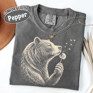 May include: A charcoal gray t-shirt featuring a graphic of a bear holding a dandelion with seeds blowing away. The shirt has a crew neckline and a Comfort Colors label at the inner neck. A thin silver chain necklace with a small round pendant rests on top.