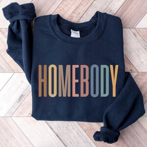 May include: Navy blue sweatshirt with the word "HOMEBODY" in a colorful, retro font across the front. The sweatshirt has a crew neck and long sleeves with ribbed cuffs. The garment is made of a soft material.