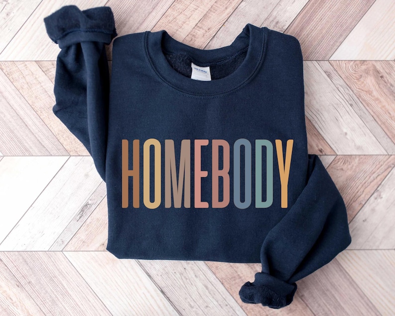 May include: Navy blue sweatshirt with the word "HOMEBODY" in a colorful, retro font across the front. The sweatshirt has a crew neck and long sleeves with ribbed cuffs. The garment is made of a soft material.