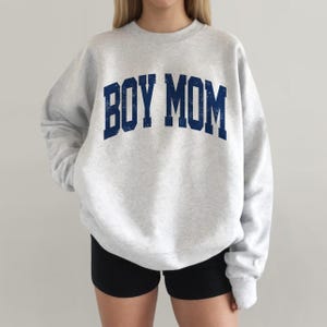 May include: A light gray sweatshirt with the text "BOY MOM" in navy blue, collegiate-style lettering. The sweatshirt is a crew neck style.