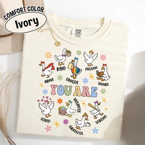 May include: Ivory-colored Comfort Colors t-shirt featuring cartoon geese with positive affirmations. The geese are depicted with various accessories and phrases like "Brave," "Kind," and "Unique." The words "YOU ARE" are prominently displayed.