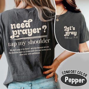 K&ouml;nnte beinhalten: Dunkelgraues T-Shirt mit dem Text "need prayer? tap my shoulder" in wei&szlig;er Retro-Schrift. Auf der R&uuml;ckseite des Shirts befindet sich auch ein Zitat. Ein kleineres Bild auf der rechten Seite zeigt "Let's pray Hebrews 4:16". Das Shirt ist mit "Comfort Color Pepper" gekennzeichnet.