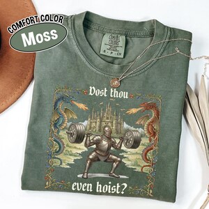 May include: Moss green t-shirt with a medieval knight lifting weights, flanked by dragons, and the text "Dost thou even hoist?" The shirt also features the text "Comfort Color Moss". A silver necklace is draped across the shirt.
