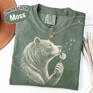 May include: A moss green short-sleeve t-shirt featuring a graphic of a bear holding a dandelion. The bear is depicted in profile, blowing on the dandelion seeds. The shirt has a crew neckline and a label that reads Comfort Colors, S P CH.