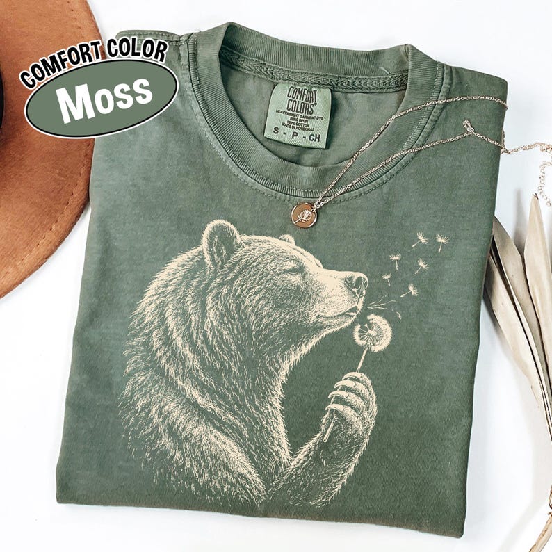 May include: A moss green short-sleeve t-shirt featuring a graphic of a bear holding a dandelion. The bear is depicted in profile, blowing on the dandelion seeds. The shirt has a crew neckline and a label that reads Comfort Colors, S P CH.