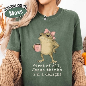 May include: A moss green t-shirt featuring a graphic of a toad wearing a pink bow and holding a pink mug with a heart. Below the toad, the text reads: first of all, Jesus thinks i'm a delight. The shirt has a crew neckline and short sleeves.