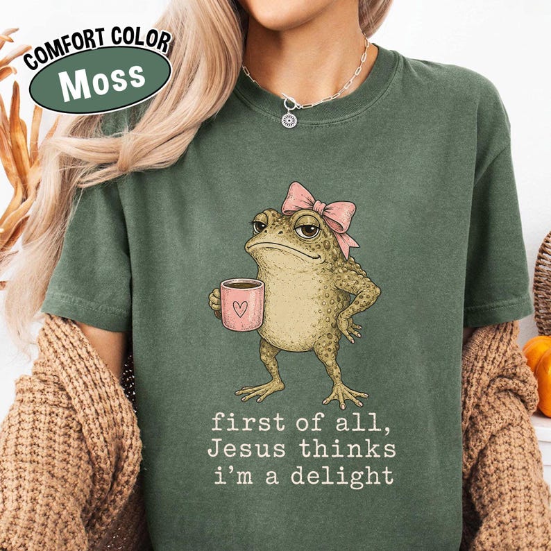 May include: A moss green t-shirt featuring a graphic of a toad wearing a pink bow and holding a pink mug with a heart. Below the toad, the text reads: first of all, Jesus thinks i'm a delight. The shirt has a crew neckline and short sleeves.