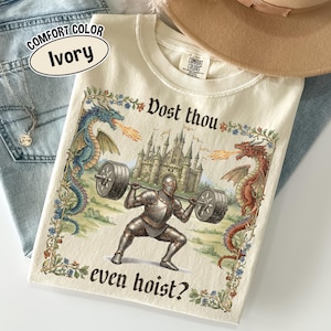 May include: Ivory-colored t-shirt with a medieval-themed graphic. The design features a knight lifting weights, flanked by dragons, with a castle in the background. Text includes "Dost thou even hoist?" and "Comfort Color Ivory".