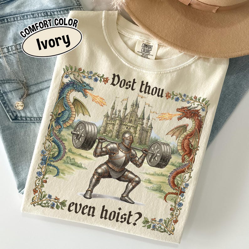 May include: Ivory-colored t-shirt with a medieval-themed graphic. The design features a knight lifting weights, flanked by dragons, with a castle in the background. Text includes "Dost thou even hoist?" and "Comfort Color Ivory".