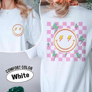 May include: White Comfort Colors t-shirt with a smiley face and lightning bolt design. The front features a small smiley face with lightning bolts, while the back has a larger design on a pink and white checkered background. The text "COMFORT COLOR White" is also visible.
