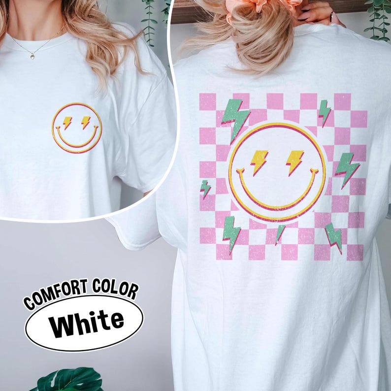 May include: White Comfort Colors t-shirt with a smiley face and lightning bolt design. The front features a small smiley face with lightning bolts, while the back has a larger design on a pink and white checkered background. The text "COMFORT COLOR White" is also visible.