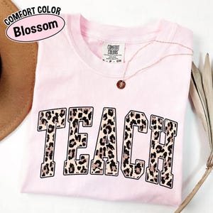 May include: A light pink Comfort Colors t-shirt with the word "TEACH" in a leopard print design. A gold necklace with a pendant is draped across the shirt. The shirt is folded and has a tag.