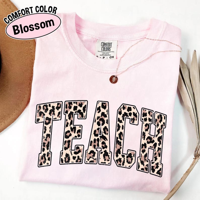 May include: A light pink Comfort Colors t-shirt with the word "TEACH" in a leopard print design. A gold necklace with a pendant is draped across the shirt. The shirt is folded and has a tag.