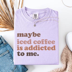 Peut inclure: T-shirt lilas pli&eacute; avec l'inscription "maybe iced coffee is addicted to me" en marron et beige. L'&eacute;tiquette Comfort Colors est visible. Un collier argent&eacute; avec un pendentif est pos&eacute; dessus. V&ecirc;tement de la collection.
