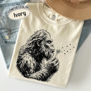 Puede incluir: Una camiseta de color marfil con una ilustraci&oacute;n gr&aacute;fica en negro de una criatura peluda soplando un diente de le&oacute;n. La prenda tiene cuello redondo y una etiqueta de la marca Comfort Colors en el interior. Un globo de texto dice Comfort Color Ivory.