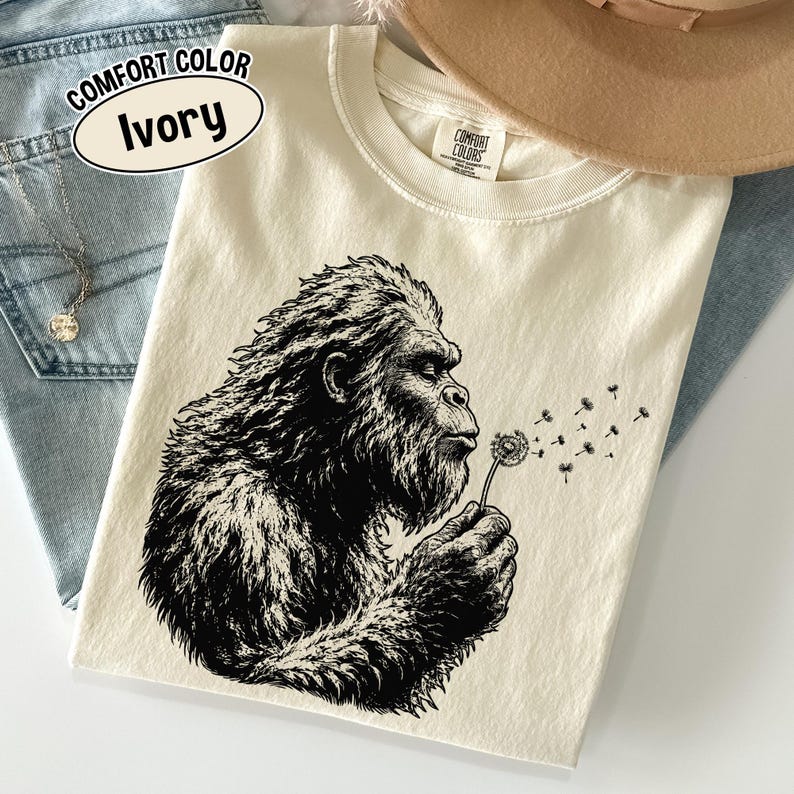 Puede incluir: Una camiseta de color marfil con una ilustraci&oacute;n gr&aacute;fica en negro de una criatura peluda soplando un diente de le&oacute;n. La prenda tiene cuello redondo y una etiqueta de la marca Comfort Colors en el interior. Un globo de texto dice Comfort Color Ivory.